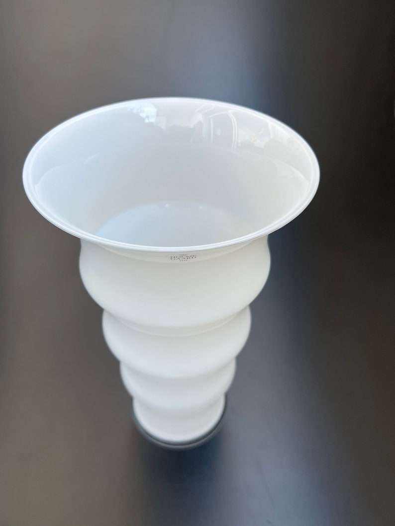 May include: White ceramic vase with a flared top and a rounded, wavy body. The vase has a black base.