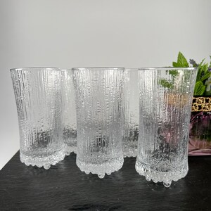 May include: A set of six clear, textured drinking glasses. The glasses have a vertical, bark-like pattern and a decorative base. The glasses are displayed on a dark surface against a neutral background. The glasses are approximately 6 inches tall.