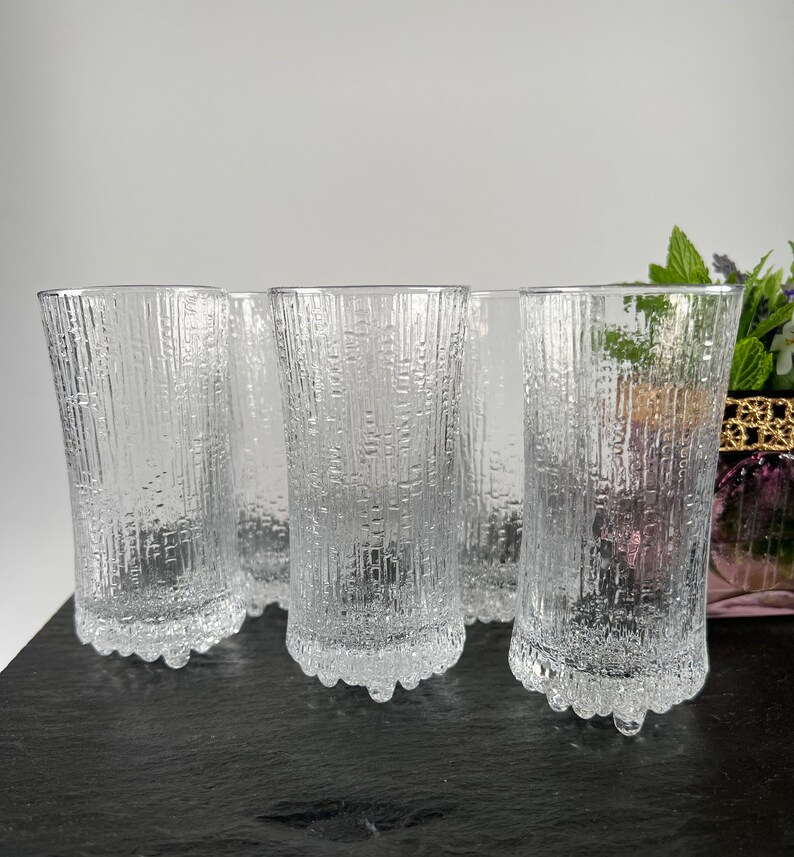 May include: A set of six clear, textured drinking glasses. The glasses have a vertical, bark-like pattern and a decorative base. The glasses are displayed on a dark surface against a neutral background. The glasses are approximately 6 inches tall.
