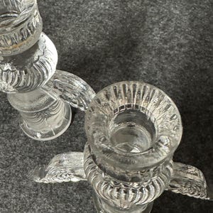 Puede incluir: Dos candelabros de vidrio transparente con un dise&ntilde;o decorativo y texturizado. Cada soporte tiene una parte superior circular para la vela y una base con un dise&ntilde;o en forma de ala. Los candelabros est&aacute;n sobre una superficie gris.