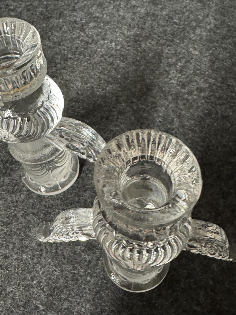Puede incluir: Dos candelabros de vidrio transparente con un dise&ntilde;o decorativo y texturizado. Cada soporte tiene una parte superior circular para la vela y una base con un dise&ntilde;o en forma de ala. Los candelabros est&aacute;n sobre una superficie gris.