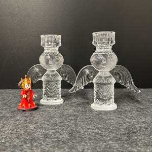 Puede incluir: Dos candelabros de &aacute;ngel de cristal transparente con alas y caras detalladas. Una peque&ntilde;a figura en rojo y dorado se encuentra a la izquierda de los candelabros. Los candelabros est&aacute;n sobre una superficie gris.