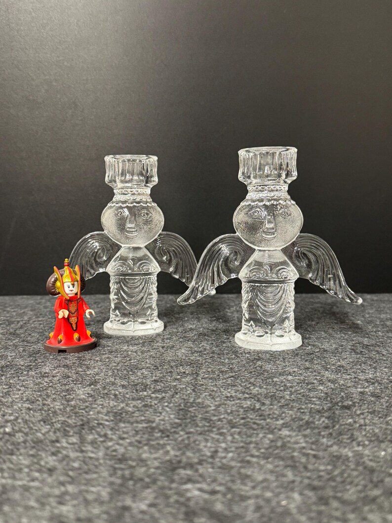 Puede incluir: Dos candelabros de &aacute;ngel de cristal transparente con alas y caras detalladas. Una peque&ntilde;a figura en rojo y dorado se encuentra a la izquierda de los candelabros. Los candelabros est&aacute;n sobre una superficie gris.