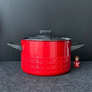 Op de afbeelding: Een felrode kookpot met een zwart deksel en handvatten. De pot heeft een gestructureerd patroon aan de buitenkant. Een klein beeldje staat rechts van de pot. De achtergrond is donkergrijs.