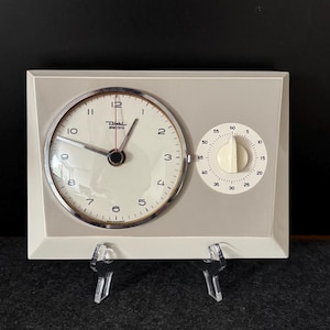 Może przedstawiać: Zegar kuchenny i minutnik Dahl electro w stylu vintage. Zegar ma białą tarczę z czarnymi cyframi i chromowaną ramką. Tarcza timera jest biała z czarnymi oznaczeniami. Urządzenie znajduje się w jasnoszarej prostokątnej ramie.