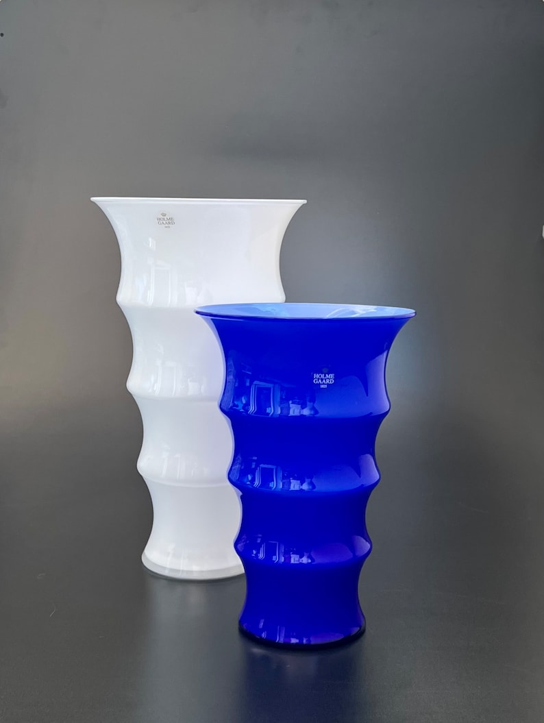 May include: Two glass vases, one white and one blue, with a ribbed design. The vases are both tall and slender, and they have a wide mouth. The white vase has the text "HOLME GARD  DK" on the side. The blue vase has the text "HOLME GARD  DK" on the side.