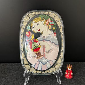 Pode incluir: Um prato decorativo oval transparente com uma mulher em um vestido segurando um soldado de brinquedo. O prato tem uma borda dourada e preta ornamentada. Uma pequena figura vermelha e dourada está ao lado do prato.