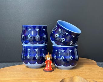 May include: Four blue ceramic cups with white starburst patterns and a small red figurine on a wooden board. The cups have a dark blue exterior and a lighter blue interior.
