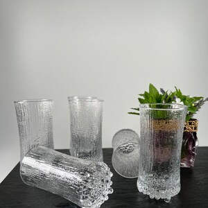 May include: A collection of clear, textured drinking glasses. The glasses have a vertical, bark-like pattern and are cylindrical in shape. Some glasses are upright, while others are lying on their sides. The glasses are on a dark surface.