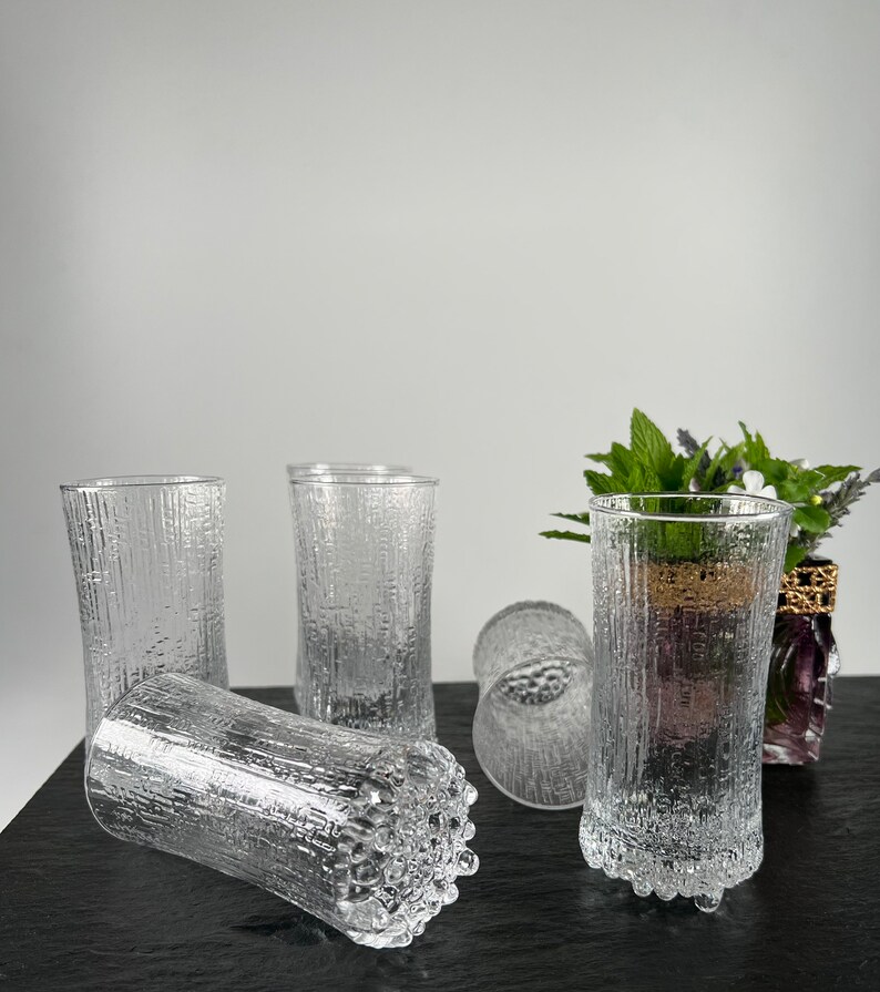 May include: A collection of clear, textured drinking glasses. The glasses have a vertical, bark-like pattern and are cylindrical in shape. Some glasses are upright, while others are lying on their sides. The glasses are on a dark surface.