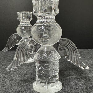 Puede incluir: Dos candelabros de &aacute;ngel de cristal transparente. Cada uno tiene una cara, alas y un portavelas. Los candelabros est&aacute;n sobre una superficie gris. Los candelabros de &aacute;ngel son decorativos.