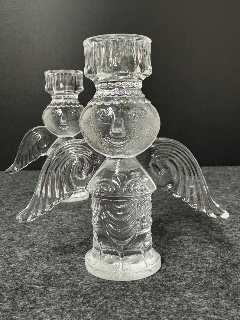Puede incluir: Dos candelabros de &aacute;ngel de cristal transparente. Cada uno tiene una cara, alas y un portavelas. Los candelabros est&aacute;n sobre una superficie gris. Los candelabros de &aacute;ngel son decorativos.