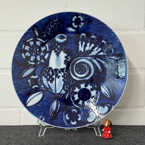 Puede incluir: Un plato decorativo redondo azul con un diseño de pájaro y flores. El plato presenta un fondo azul oscuro con detalles en blanco y azul claro. Una pequeña figura roja se encuentra frente al plato.