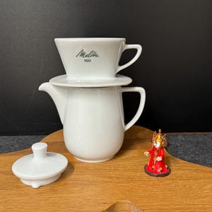 Puede incluir: Un juego de cafetera de cerámica blanca con un filtro Melitta 100, una tetera y una tapa. Una pequeña figura roja y dorada está sobre una tabla de madera.
