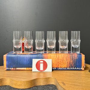 May include: Set of six clear glass shot glasses with a textured, bumpy surface. The glasses are arranged in a row on a blue and orange box with the text "DESIGN TAPIO WIRKKALA".
