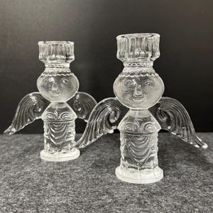 Puede incluir: Dos candelabros de &aacute;ngel de cristal transparente. Cada &aacute;ngel tiene una cara, alas y un cuerpo detallado. Los candelabros est&aacute;n sobre una superficie gris con un fondo oscuro. Los &aacute;ngeles son un objeto decorativo.
