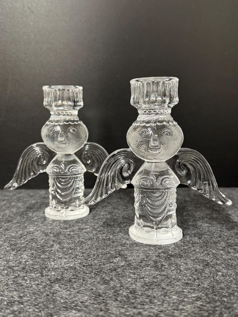 Puede incluir: Dos candelabros de &aacute;ngel de cristal transparente. Cada &aacute;ngel tiene una cara, alas y un cuerpo detallado. Los candelabros est&aacute;n sobre una superficie gris con un fondo oscuro. Los &aacute;ngeles son un objeto decorativo.