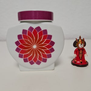 May include: White ceramic jar with a purple lid and a red and purple flower design. A small red and gold figurine stands next to the jar.