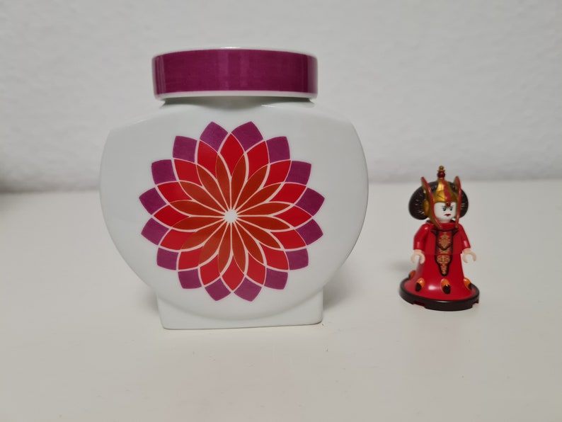 May include: White ceramic jar with a purple lid and a red and purple flower design. A small red and gold figurine stands next to the jar.
