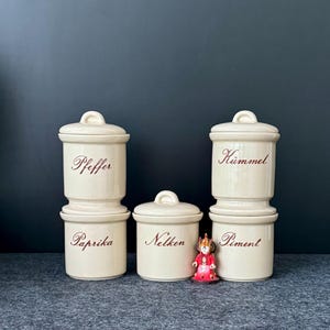 May include: A collection of off-white ceramic spice jars with lids, labeled "Pfeffer", "Paprika", "Nelken", "Kümmel", and "Piment" in brown script. A small, decorative figurine stands in front of the jars. The jars are arranged on a gray surface against a dark background.