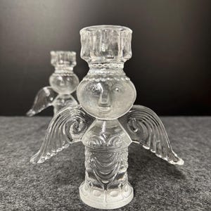 Puede incluir: Dos candelabros de &aacute;ngel de cristal transparente. Cada &aacute;ngel tiene una cara, alas y un portavelas. Los candelabros est&aacute;n sobre una superficie gris con un fondo oscuro. Los &aacute;ngeles est&aacute;n detallados con dise&ntilde;os grabados.