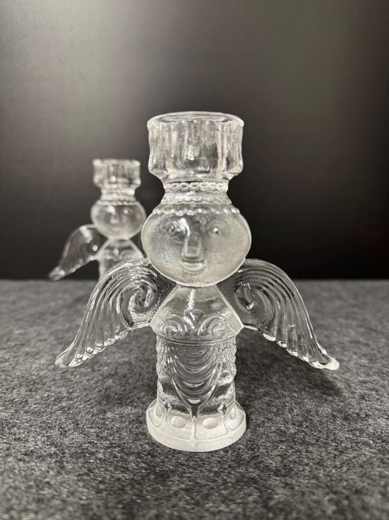 Puede incluir: Dos candelabros de &aacute;ngel de cristal transparente. Cada &aacute;ngel tiene una cara, alas y un portavelas. Los candelabros est&aacute;n sobre una superficie gris con un fondo oscuro. Los &aacute;ngeles est&aacute;n detallados con dise&ntilde;os grabados.