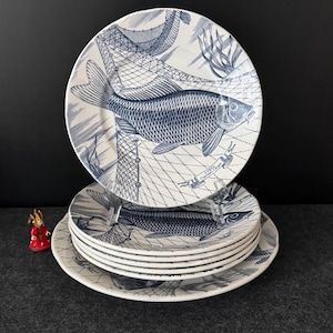 Könnte beinhalten: Ein Satz weißer Keramikplatten mit blauem Fischdesign. Die Teller zeigen eine detaillierte Illustration eines Fisches, eines Fischernetzes und Wasserpflanzen. Die Teller sind übereinander gestapelt und präsentieren das Design. Im Hintergrund ist eine kleine Figur zu sehen.