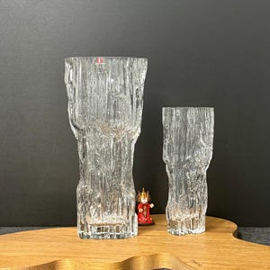 Könnte beinhalten: Zwei klare Glasvasen mit einem strukturierten, rindenartigen Design. Die größere Vase ist etwa 25 cm hoch, die kleinere etwa 15 cm. Sie stehen auf einem Holzbrett mit organischer Form. Zwischen den Vasen befindet sich eine kleine Figur.