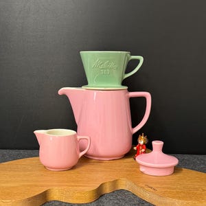 Op de afbeelding: Een roze keramische koffiepot, melkkan en deksel worden gepresenteerd op een houten oppervlak. Een groene keramische koffiefilter met de tekst "Melitta 101" staat bovenop de pot. Er is ook een klein beeldje aanwezig.