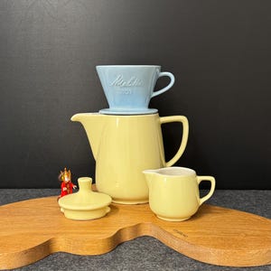 Op de afbeelding: Een gele keramische koffiepot, melkkan en deksel staan op een houten serveerplank. Een lichtblauw Melitta koffiefilter staat bovenop de pot. Er staat ook een klein beeldje op de plank.