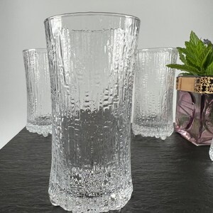 May include: Clear, textured drinking glasses. The glasses have a vertical, bark-like pattern. The base of the glasses have a rounded, textured design. The image shows four glasses, with one in the foreground and three in the background.