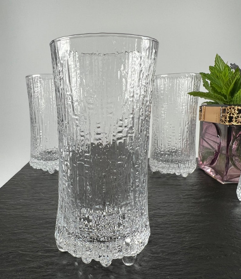May include: Clear, textured drinking glasses. The glasses have a vertical, bark-like pattern. The base of the glasses have a rounded, textured design. The image shows four glasses, with one in the foreground and three in the background.