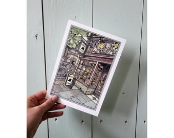 Könnte beinhalten: Ein Kunstdruck, der ein Hutgeschäft mit einem Schild "Hats & Caps" zeigt. Das Geschäft hat eine detaillierte Fassade mit einer Hutauslage. Eine Person geht auf dem Bürgersteig vor dem Geschäft. Der Druck wird von einer Hand gehalten.