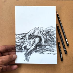 May include: A charcoal drawing on white paper depicting a figure in a reclining pose with arms extended overhead. The artwork features expressive shading and dark textures. Three black drawing pencils are positioned to the right of the paper.