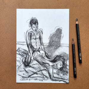 May include: A charcoal drawing on white paper depicting two figures in a reclining pose, with one figure positioned behind the other. The artwork features expressive, loose graphite strokes and a signature at the bottom. Two black artist pencils lie beside the paper.
