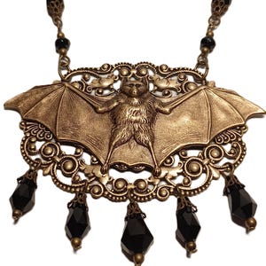 Könnte beinhalten: Eine bronzefarbene Fledermaus-Anhänger-Halskette mit aufwendigen Filigranarbeiten. Die Fledermaus hat ausgebreitete Flügel und befindet sich in einer dekorativen Platte. Schwarze, facettierte Perlen hängen von der Unterseite des Anhängers.