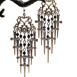 May include: A pair of ornate, bronze-toned earrings with a gothic aesthetic. Each earring features a filigree top, black bead accents, and multiple dangling dagger charms. The earrings are suspended from a black, textured branch against a white background.