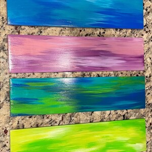 May include: Six wooden signs painted in different colors, including pink, blue, purple, green, lime green, and light blue. Each sign has a horizontal brushstroke pattern.