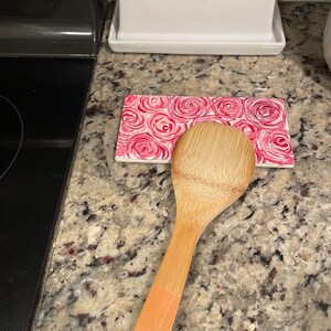 May include: A wooden spoon with a light brown handle rests on a pink and white floral patterned trivet. The trivet is on a granite countertop.