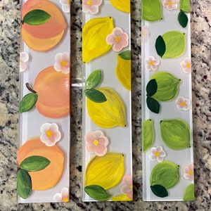 May include: Three rectangular glass tiles with painted fruit and flower designs. The first tile features peaches, the second tile features lemons, and the third tile features limes. All three tiles have a white background with pink flowers.