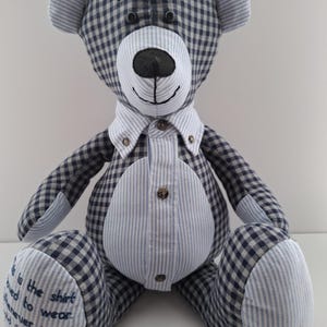 Pode incluir: Um urso de peluche xadrez azul e branco vestindo uma camisa listrada azul e branca. O urso está sentado com as patas à sua frente. As patas do urso têm um texto bordado que diz "This is the shirt I loved to wear whenever you would know I am there."