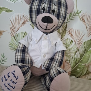 Puede incluir: Un oso de peluche hecho de tela a cuadros azul marino, blanca y beige con detalles en beige sólido en las orejas, el hocico y las patas. El oso lleva una camisa blanca abotonada y tiene el texto "Don't be sad and don't be lonely" bordado en su pie izquierdo.