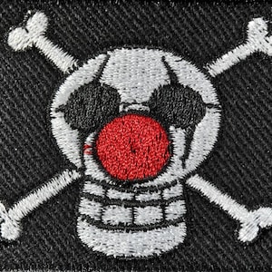 May include: Embroidered patch featuring a skull with crossbones, a red nose, and black eye patches. The skull is white with black accents, set against a black fabric background. The patch has a stitched design.
