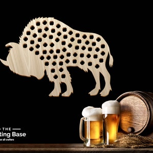 Puede incluir: Abrebotellas de bisonte de madera con 100 agujeros para tapas de botellas. El bisonte está de pie sobre una superficie de madera con dos vasos de cerveza y un barril en el fondo.