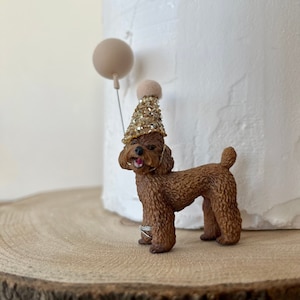 May include: A brown poodle figurine wearing a gold sequined party hat and two silver rings. A beige balloon on a wire is attached to the figurine. The figurine is on a wooden surface, with a white textured background.