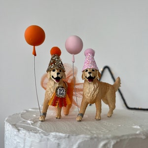 May include: Two golden retriever cake toppers, each wearing a party hat and holding a balloon. One dog has a sequined hat and a sash with the number 12. The balloons are orange and pink.