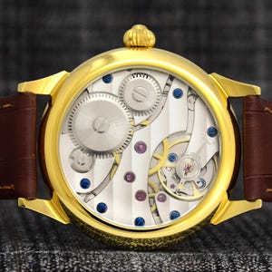 May include: A gold-toned wristwatch with a brown leather band. The watch face reveals intricate mechanical components, including gears, screws, and jewels. The watch has a round face and a textured, striped background.