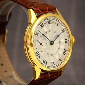 May include: A gold-toned wristwatch with a light-colored face and Roman numeral hour markers. The watch features a brown leather band and a smaller dial. The watch face reads "ROMAN MILLER".