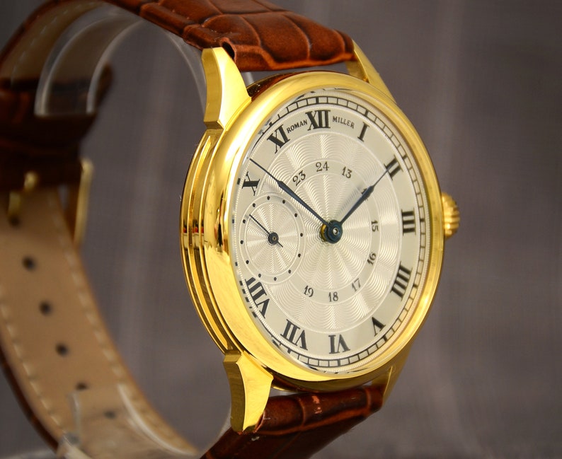 May include: A gold-toned wristwatch with a light-colored face and Roman numeral hour markers. The watch features a brown leather band and a smaller dial. The watch face reads "ROMAN MILLER".