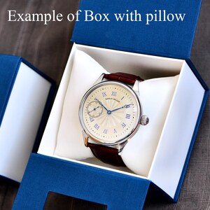May include: A silver wristwatch with a brown leather band rests on a white pillow inside a blue and white box. The watch face is cream-colored with blue hands and Roman numerals. The box is open, with the lid propped up.